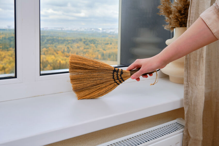 A woman sweeps dust with a broom from a windowsill in a home living room